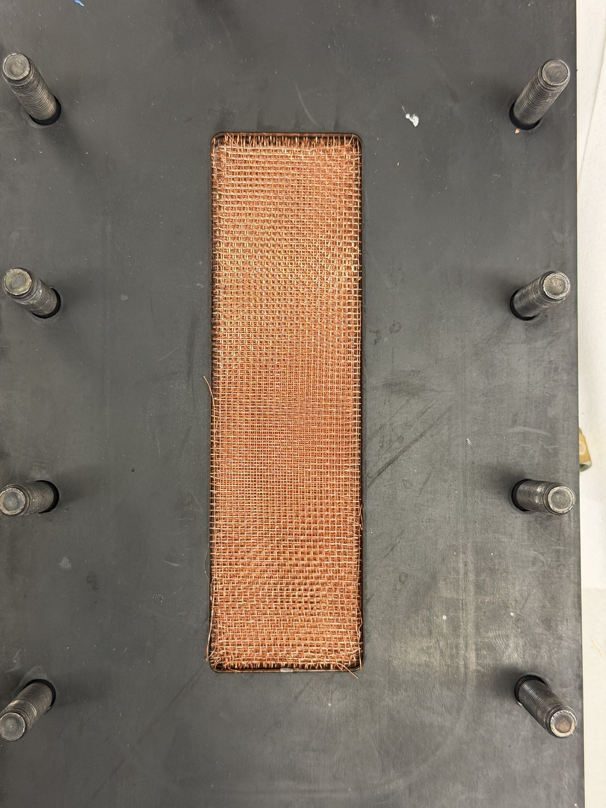 Top-down view of a machined module plate with a copper wire mesh insert set into a rectangular slot, surrounded by threaded mounting studs.