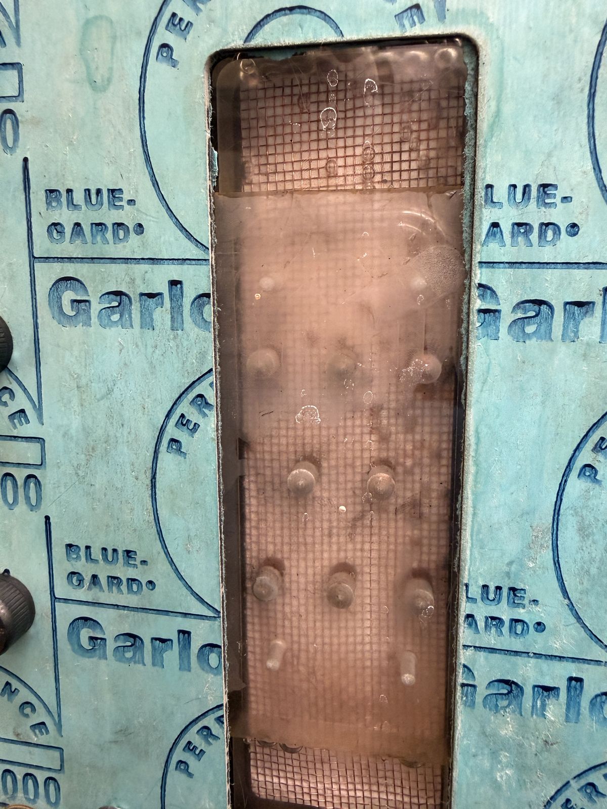 Close-up through a Garlock gasket sheet window showing a finned module insert with a row of pin-fin contacts visible inside the housing cavity.