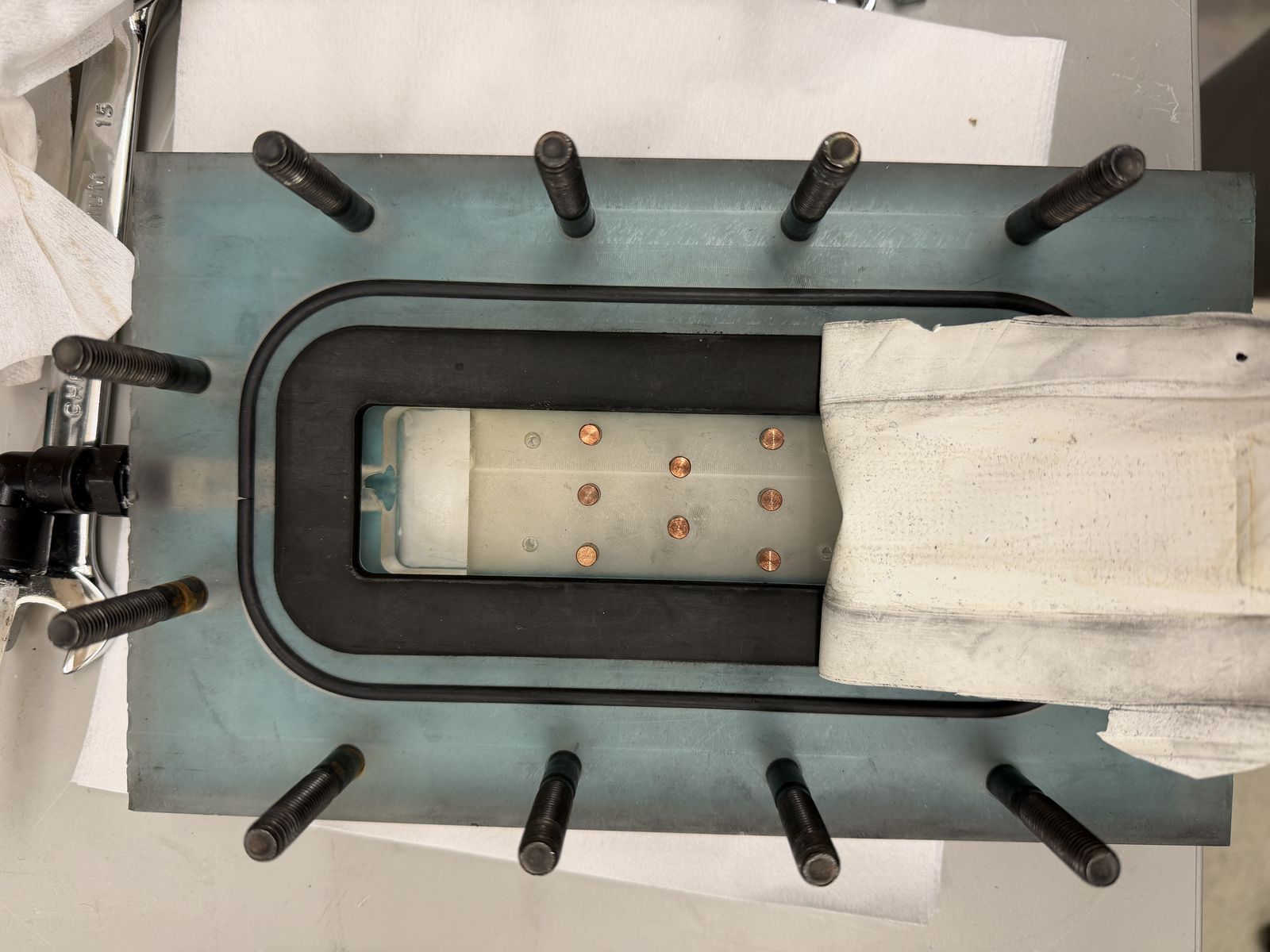 Overhead view of the module housing plate bolted down with twelve studs, showing a rounded rectangular gasket seal around a membrane window with copper pin contacts, a gloved hand and wrench visible at the edge.